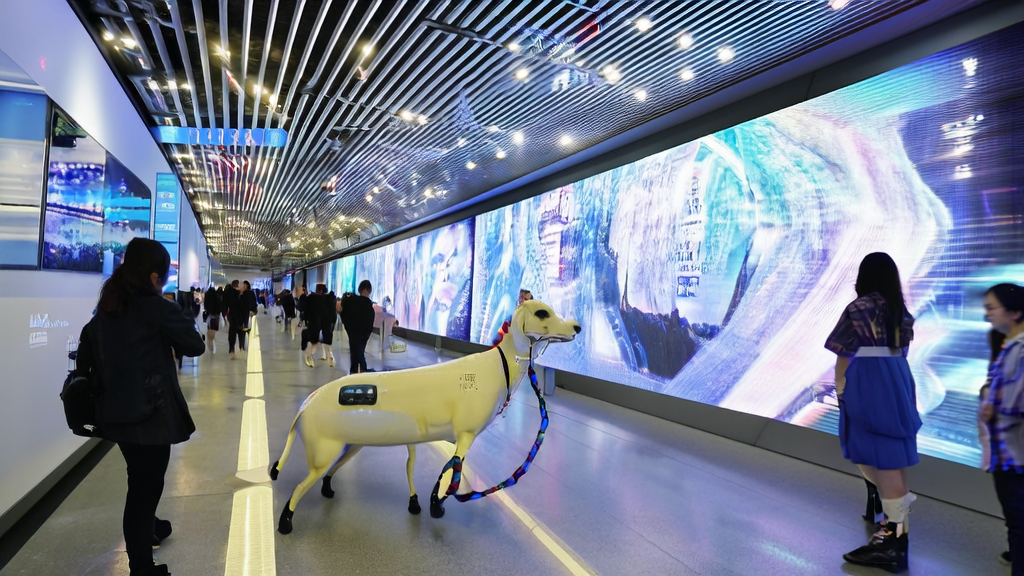 In 2025, at a Shenzhen subway station, an AI-powered guide dog “Xiaosuan“ assists a visually impaired person while AI-optimized cross-border e-commerce ads are displayed around.